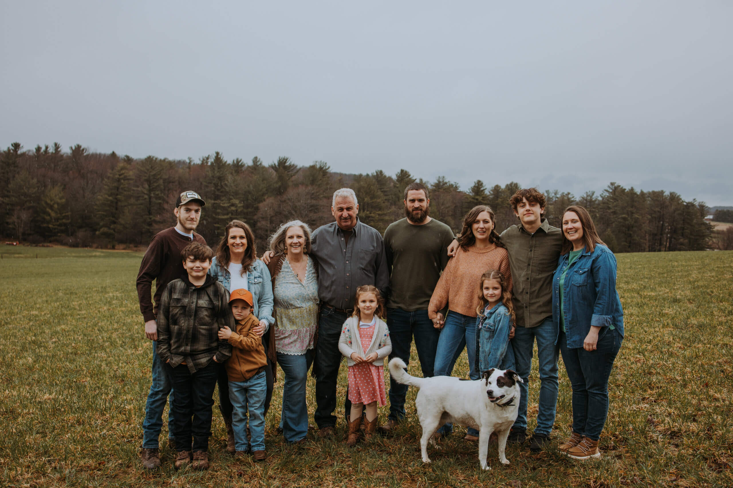 family standing together in a field with a dog in the front and trees in the background. 